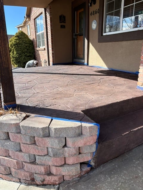 Patio under construction with stamped concrete surface, brick border edging, and protective black fabric, adjacent to brown house entrance