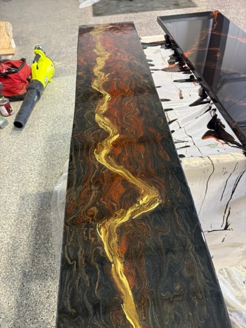 Long dark stone countertop with gold resin inlay being inspected at outdoor warehouse, with worker in yellow safety vest nearby