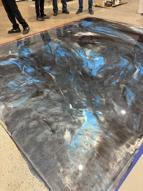 Large dark fabric or tarp with blue spray paint markings on concrete ground surrounded by people wearing dark clothing