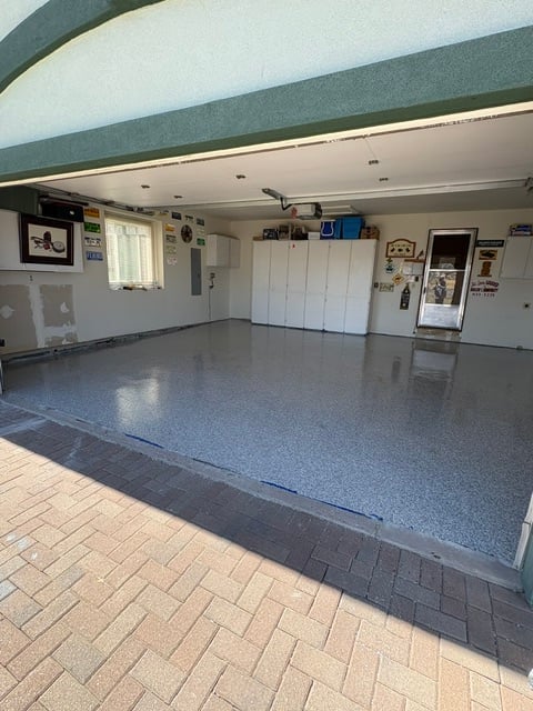 Empty residential garage with white cabinets, gray epoxy floor, and brick entry, lit by ceiling fixtures