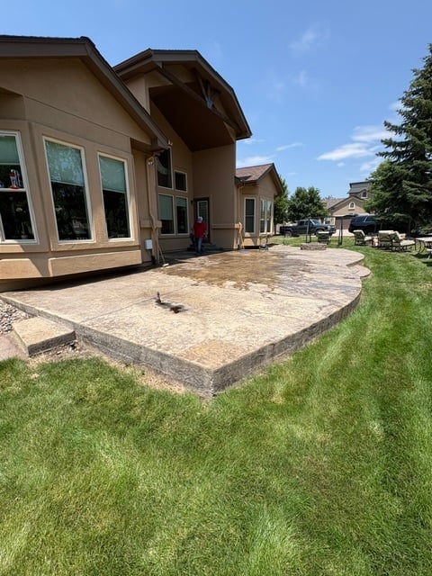 Backyard view of beige house with stone patio, manicured lawn, and neighboring homes under clear blue sky