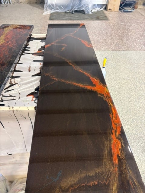 Large dark canvas with orange and rust-colored abstract brushstrokes, laid flat on concrete floor in workshop setting