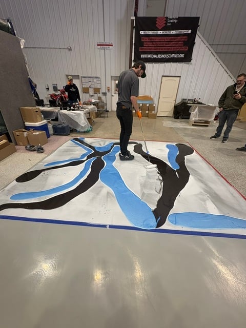 Artist painting a large abstract mural with blue and black strokes on white canvas in an industrial warehouse space with onlookers
