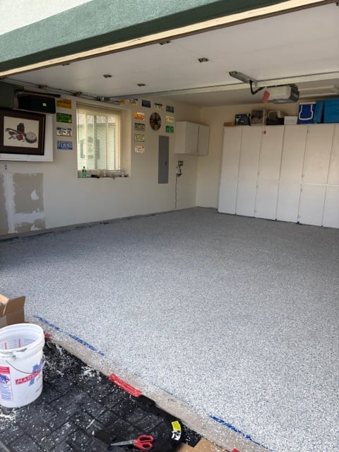 Empty garage interior with gray speckled flooring, white wall cabinets, window, overhead lighting, and storage items on upper shelves