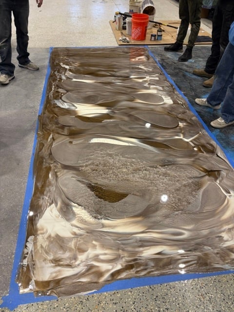 Large rectangular sandbox or sensory table with brown sand and wave patterns, bordered by blue tape, with people standing around it