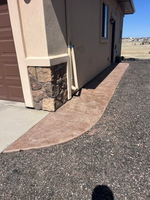 Red brick pathway leading to a modern house entrance with stone chimney and gravel driveway on a clear day