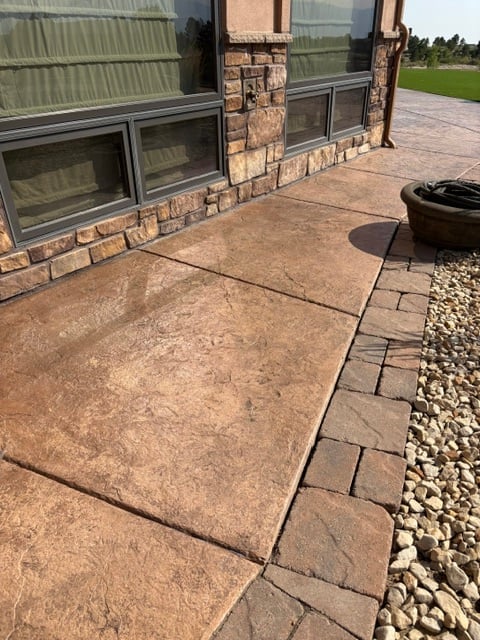 Modern patio with brown concrete pavers and brick border beside stone and glass residential building with green lawn beyond
