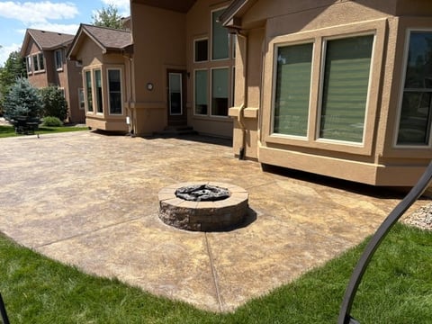 Backyard patio with stone fire pit surrounded by tan-colored pavers, bordered by green lawn and modern home with large windows