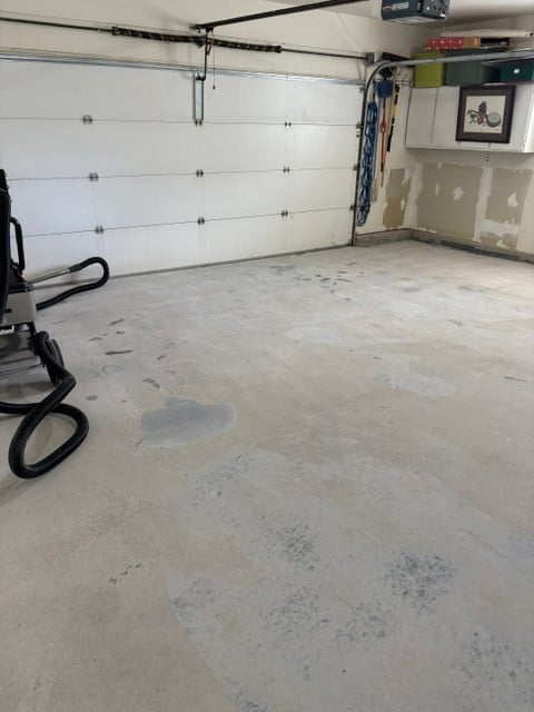 Empty garage interior with white overhead door, concrete floor, garage door opener, and black hose coiled on left side