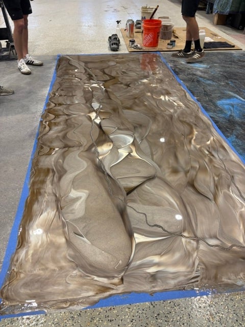 Large canvas with abstract swirled paint patterns bordered by blue tape on concrete floor, two people standing nearby with supplies
