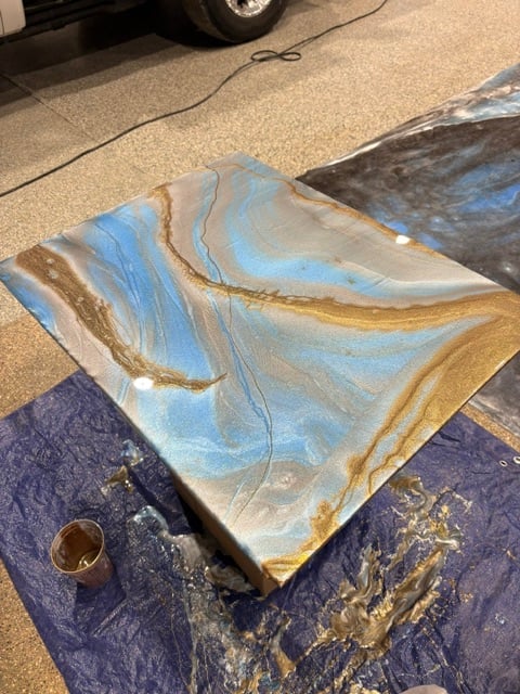 Marbled paint art piece with swirled blue, tan, and gold colors drying on blue tarp in driveway